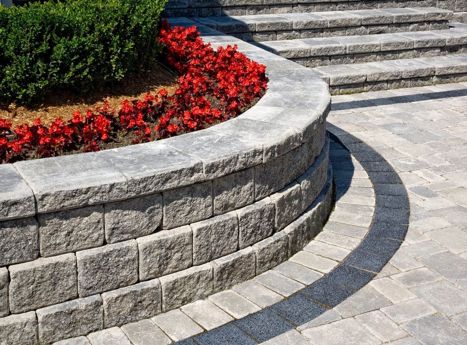 Picture of a masonry lawn made up of stone right next to entrance stairs containing red flowers and green plants